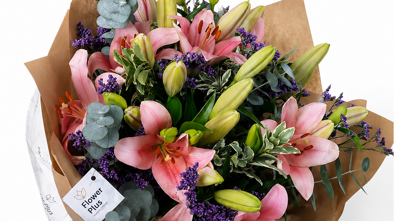 A vibrant floral bouquet wrapped in brown kraft paper and a white ribbon, featuring pink lilies, purple lavender, green buds, and eucalyptus on a white background.